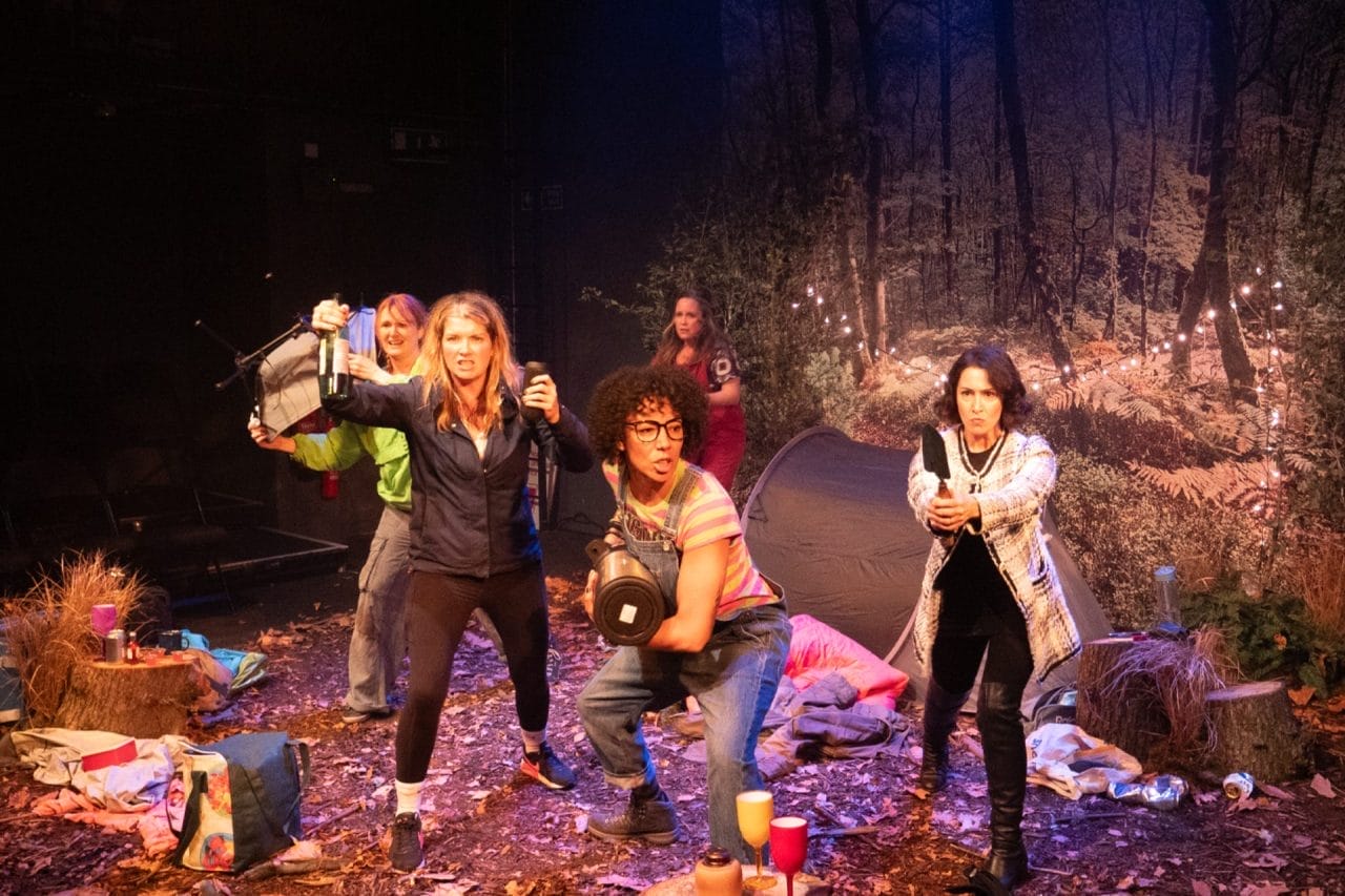 All 5 women are turned towards the camera but looking beyond it. They hold items up as if ready to fight. Stacy has a water heater, Rendah has a trowel, and Ciara has a bottle of wine. Scarlett and Sarah are in the background. Around them is a forest scene with leaves on the floor and two tree stumps with plastic wine glasses on. Behind them is a background of a forest, a tent and white fairy lights.